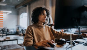 Woman with dark hair in a brown jumper operating a PC using a mouse.